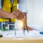 A male architect works at desk with house model toy and A3 paper blueprints, construction real estate law, zoning, permits, and resolving property disputes through arbitration and litigation.