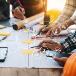 Engineers collaborate on blueprints and project models during a teamwork meeting in an office setting. The team, including Asian industry professionals, works together on building plans.