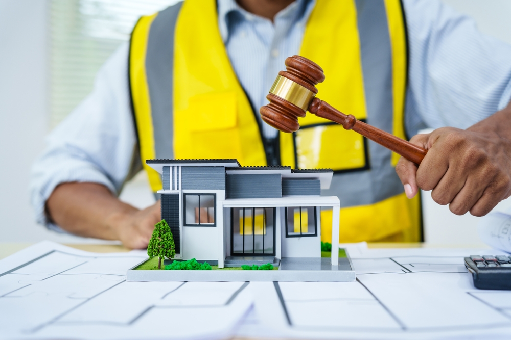 Asian male architect working at a desk with a house model toy, A3 paper blueprints, and documents, focusing on construction, real estate law, zoning, and property dispute resolution.