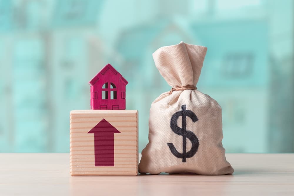 Money bag with dollar symbol and house model on a wooden cube with an upward arrow. Concept of rising real estate and rent costs.